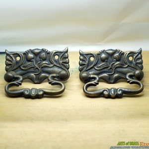 May include: Two antique brass drawer pulls with ornate floral designs and a curved handle. The pulls have a distressed finish and are perfect for adding a vintage touch to furniture.