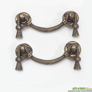 Op de afbeelding: Twee antieke messing kastgrepen met een touwontwerp. De grepen hebben een gebogen vorm en zijn aan elk uiteinde versierd met een knoop.