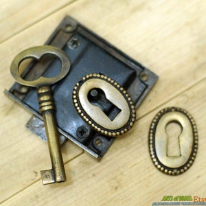 May include: A vintage brass keyhole escutcheon with a decorative beaded edge and a matching key. The escutcheon is attached to a black metal plate.