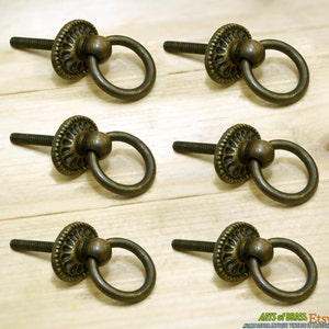 May include: Six antique brass ring pulls with a decorative floral design and a round knob. The pulls have a screw on the back for attaching to a surface.