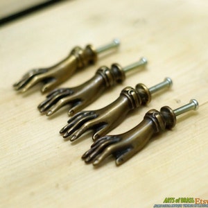 Könnte beinhalten: Vier antike Messing-Schrankknöpfe in Handform. Die Knöpfe haben einen Vintage-Look und sind perfekt, um jedem Zuhause einen Hauch von Eleganz zu verleihen.