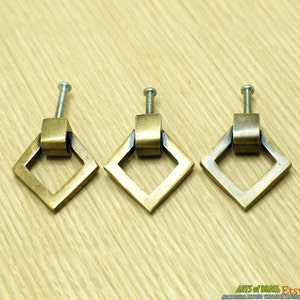 May include: Three antique brass cabinet knobs with a square shape and a ring pull. The knobs have a brushed finish and are mounted on a wooden surface.