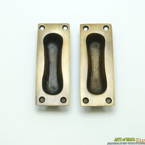 May include: Two antique brass cabinet door pulls with a rectangular shape and a recessed oval handle. The pulls have a slightly aged finish.