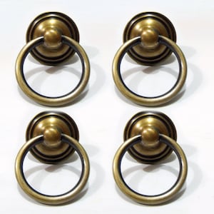 May include: Four antique gold-toned ring cabinet pulls with a black accent ring. The pulls have a round, raised centre knob.