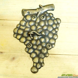 May include: A brass-toned door handle shaped like a bunch of grapes. The handle is split in two halves, with a curved latch in the center. The grapes have a textured, bumpy surface. The handle is mounted on a light-colored wooden surface.
