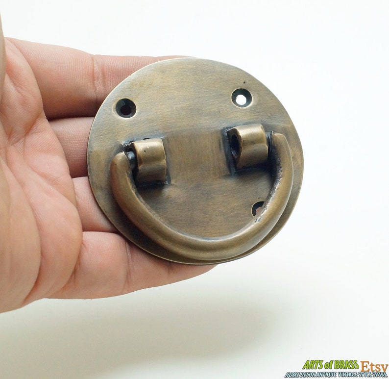 Puede incluir: Un tirador de caj&oacute;n redondo de lat&oacute;n con un asa curva. El tirador tiene un acabado desgastado y mide aproximadamente 6,5 cm de di&aacute;metro.