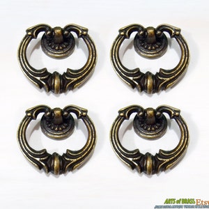 Puede incluir: Cuatro tiradores de cajón antiguos de latón con un asa en forma de anillo y detalles ornamentados. Los tiradores tienen un acabado oscuro y envejecido, dispuestos sobre un fondo blanco. Ideales para la restauración de muebles.