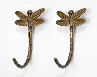 May include: Two antique brass dragonfly hooks with a curled tail. The hooks are mounted on a white wall.