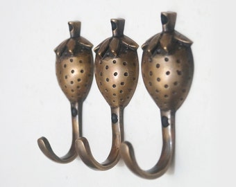 Può includere: Tre ganci in ottone a forma di fragole con una texture a pois. I ganci sono montati su una parete bianca.