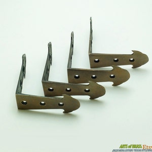 May include: Four antique brass corner brackets with a decorative arrowhead design. Each bracket has five holes for attaching to a surface.