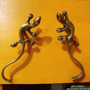 Op de afbeelding: Twee antieke messing haken in de vorm van een hagedis. De haken zijn in de vorm van een hagedis met een gekrulde staart. De haken bevinden zich op een gele achtergrond.