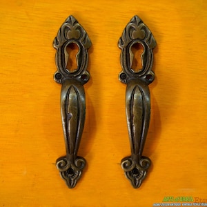 May include: Two antique brass door handles with ornate designs and keyholes. The handles are a dark gold color and have a vintage style.