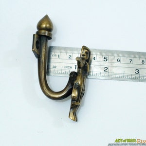 May include: Brass hook with a decorative finial and a figure of a person. The hook is 5 cm long.