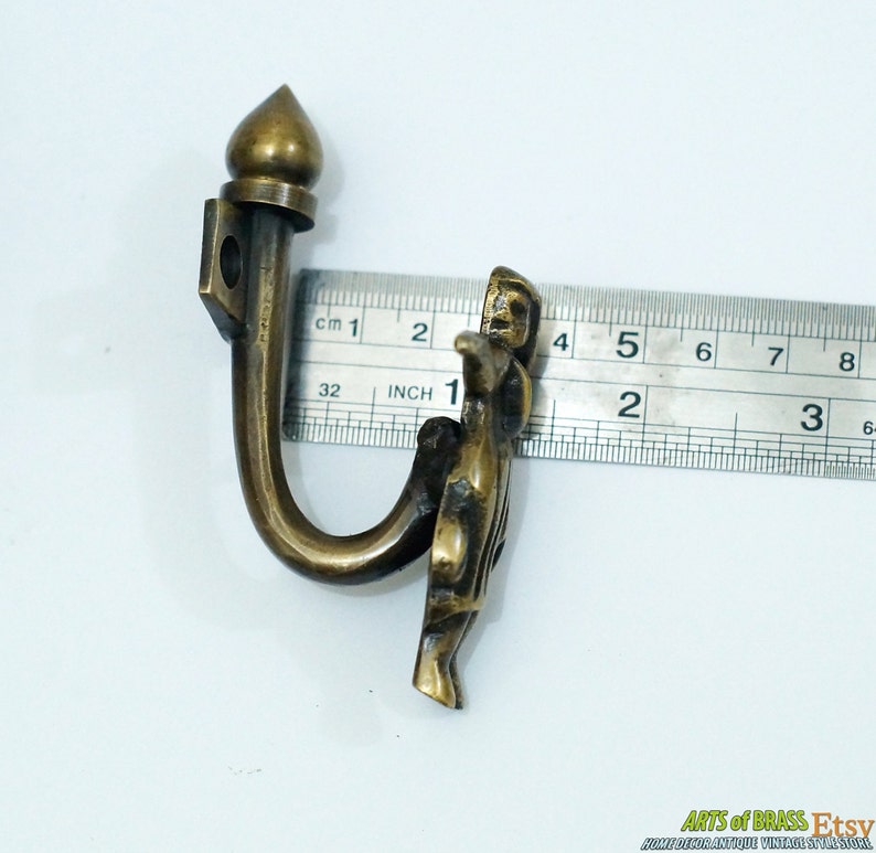 May include: Brass hook with a decorative finial and a figure of a person. The hook is 5 cm long.