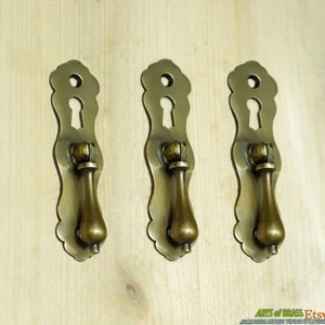 Op de afbeelding: Drie antieke messing lade handgrepen. Elke handgreep heeft een sleutelgatontwerp aan de bovenkant, een decoratieve achterplaat en een druppelvormige handgreep. Gemonteerd op een licht houten oppervlak.