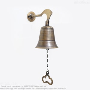 May include: A brass bell with a decorative design hangs from a wall-mounted bracket. The bell has a chain with a key-shaped pull.