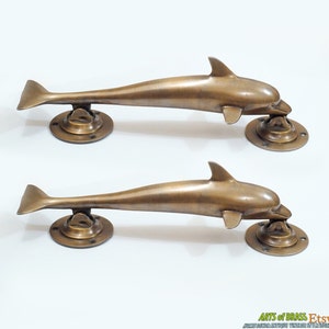 Könnte beinhalten: Zwei antike Messing-Türgriffe in Delfinform. Die Griffe sind wie Delfine geformt, mit nach oben gekrümmten Schwänzen. Die Griffe sind auf runden Messingplatten montiert.