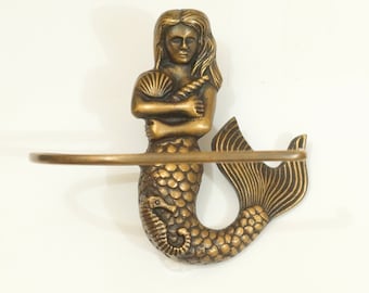 May include: A gold-toned mermaid wall shelf with a decorative seashell in her hand. The shelf is made of metal and has a curved design.