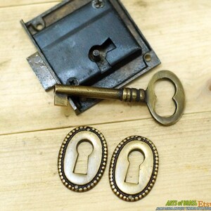 May include: A vintage brass keyhole escutcheon set with two oval keyhole covers, a brass key, and a brass lock with a black metal faceplate. The set is on a wooden surface.