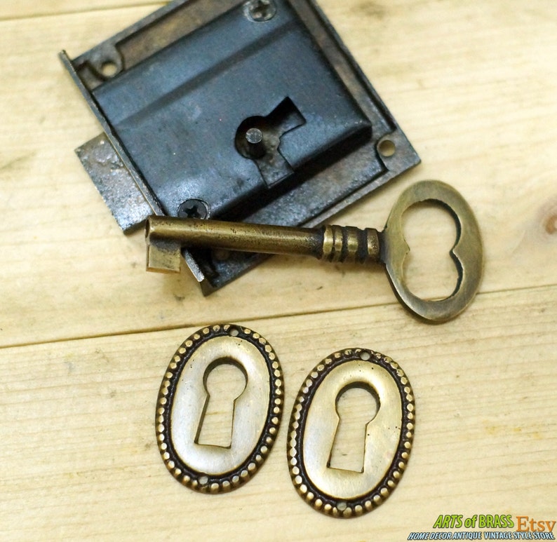 May include: A vintage brass keyhole escutcheon set with two oval keyhole covers, a brass key, and a brass lock with a black metal faceplate. The set is on a wooden surface.