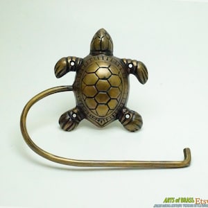 Könnte beinhalten: Ein Toilettenpapierhalter aus Messing in Form einer Schildkröte. Die Schildkröte hat einen detaillierten Panzer und ist an einem gebogenen Arm befestigt.