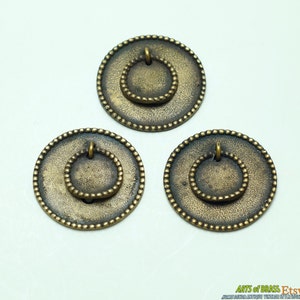 Pode incluir: Três puxadores de gaveta antigos em latão com um design circular. Cada puxador apresenta um círculo externo grande e texturizado com um círculo interno menor e com contas e uma alça de anel. Os puxadores têm uma aparência vintage e envelhecida.