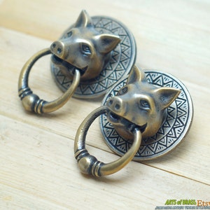 Może przedstawiać: Dwa antyczne mosiężne gałki drzwiowe w kształcie świni z ozdobnymi, okrągłymi podstawami. Każda gałka ma szczegółowo wykonaną głowę świni z uchwytem w kształcie pierścienia.