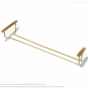 May include: A gold-colored metal wine glass rack. The rack has two horizontal bars and two rectangular end pieces. The rack is designed to hold wine glasses. The text "artsofbrass" is visible in the image.
