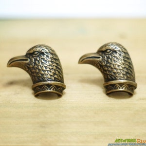 May include: Two antique brass eagle head finials with detailed feather patterns and a decorative border. The finials are a dark gold colour and have a slightly aged appearance.