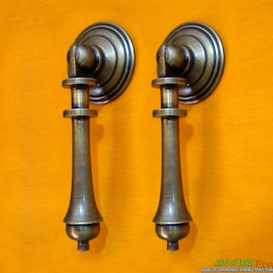 Könnte beinhalten: Zwei antike Messing-Schrankgriffe mit abgerundetem, verziertem Design. Die Griffe haben einen langen, schlanken Griff mit abgerundetem Kopf und einem dekorativen Sockel.