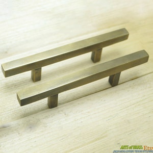 May include: Two brass cabinet pulls with a rectangular shape and a straight bar design. The pulls are shown on a light wood surface.