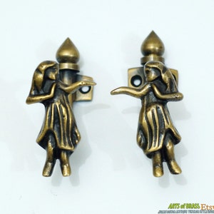 May include: A pair of antique brass door handles with a decorative design of a woman in a dress. The handles are in the shape of a woman with her arms outstretched. The handles are mounted on a square base with two screws.