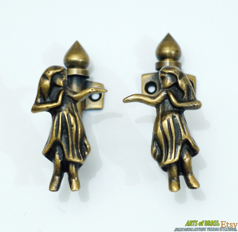 May include: A pair of antique brass door handles with a decorative design of a woman in a dress. The handles are in the shape of a woman with her arms outstretched. The handles are mounted on a square base with two screws.