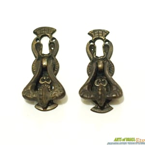 以下が含まれることがあります： 装飾的なデザインが施されたアンティークな真鍮製の引き出しノブが2つ。それぞれに鍵穴とリング状のハンドルが付いています。