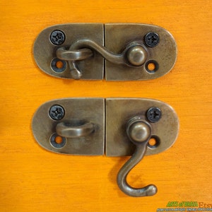 May include: Close-up of two antique brass latches with a hook-and-eye closure, each secured with two screws. The latches are mounted on a bright orange wooden surface, showcasing a vintage aesthetic.