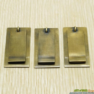 Könnte beinhalten: Drei rechteckige Anhänger aus Messing mit einem geschichteten Design. Jeder Anhänger hat ein kleineres, erhabenes Rechteck, das an einem größeren befestigt ist, mit einer kleinen Schlaufe oben zum Aufhängen. Das Messing hat eine warme, gealterte Patina.