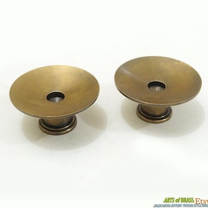 May include: Two antique brass cabinet knobs with a wide, shallow, round top and a smaller, round base. The knobs have a smooth, polished finish.