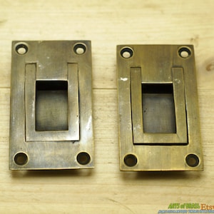 May include: Two antique brass drawer pulls with a rectangular shape and a recessed handle. The pulls have a distressed finish and four small round holes on the edges.