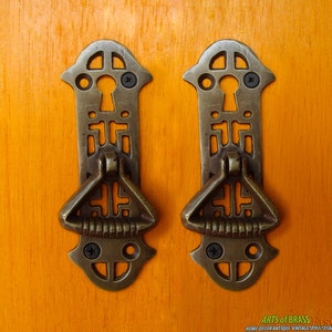 May include: Two antique brass cabinet door pulls with a keyhole design and a decorative ring handle. The pulls are mounted on a wooden surface.