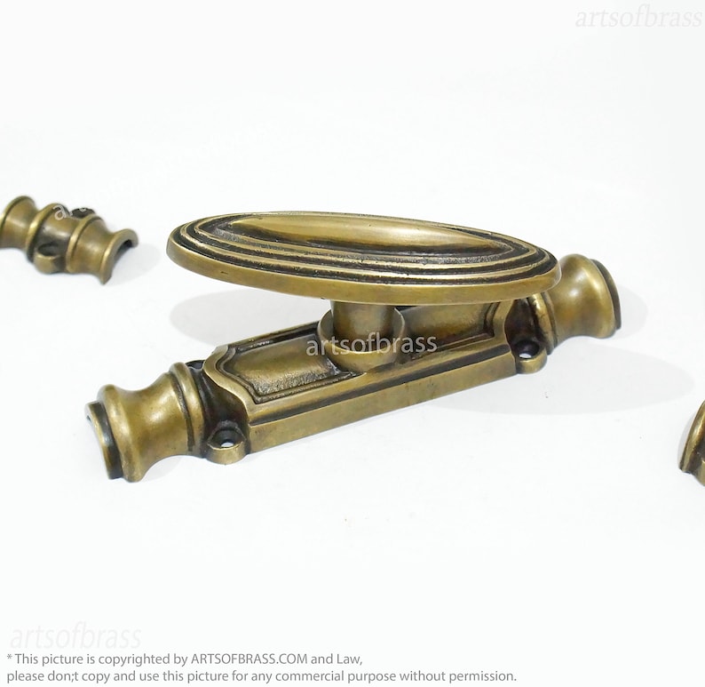 May include: A brass window latch with an oval handle and decorative details. The latch is in a polished brass finish.