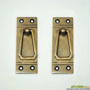 Puede incluir: Dos tiradores de puerta de armario de latón antiguo con forma rectangular y un pomo redondeado y elevado. Los tiradores tienen un borde biselado decorativo y una abertura rectangular simple.