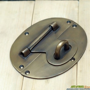 May include: A brass oval-shaped latch with a sliding bolt and a ring handle. The latch has a distressed finish and is suitable for use on a cabinet, chest, or door.
