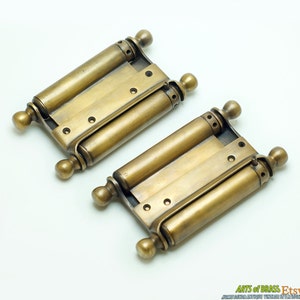 May include: Two antique brass double-action door hinges with a scroll design. The hinges are in excellent condition and have a beautiful patina.