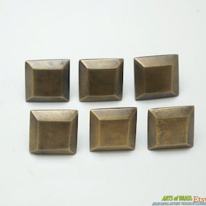 Pode incluir: Seis cabeças de prego decorativas quadradas de latão com um centro ligeiramente elevado. As cabeças de prego estão dispostas em duas filas de três.