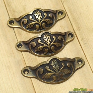 Può includere: Tre tiretti per cassetti in ottone antico con un design floreale. I tiretti hanno la forma di un semicerchio e hanno un bordo decorativo.