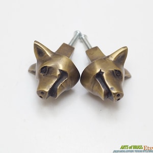 Może przedstawiać: Dwa mosiężne gałki do szafek w kształcie głowy lisa ze śrubami. Gałki są szczegółowe z cechami lisa, w tym spiczastymi uszami, pyskiem i oczami. Gałki mają ciepły, antyczny złoty kolor.