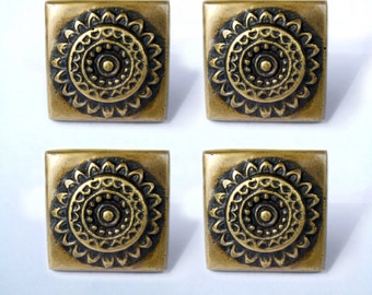 May include: Four square brass knobs with a decorative, circular, mandala-like design. The knobs are a dark gold color and have a raised, textured design.