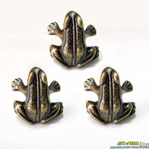 May include: Three antique brass frog drawer pulls. Each pull is detailed with a textured finish and features a frog sitting with its legs outstretched.