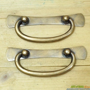 May include: Two antique brass drawer pulls with a curved, rounded handle and a flat, rectangular base. The pulls have a worn, aged finish.