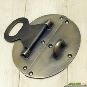 May include: A brass oval-shaped door latch with a keyhole and a hook. The latch has two hinges and is designed to be mounted on a door.